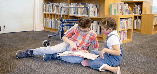 An older boy and younger girl wearing face masks sitting on the carpet in the children’s area of the library. They are looking at an open book in their laps and the boy is pointing to something in the book. There is a walker behind him.
