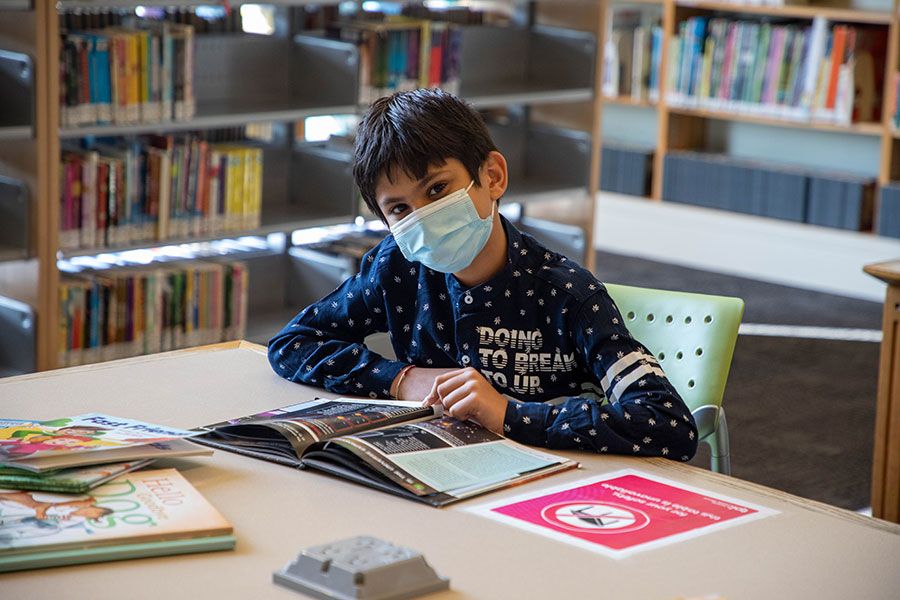 A boy sitting at a table in the library in front of an open book. He is wearing a face mask and looking at the camera. 