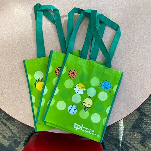 Three bright green tote bags on a table, laid flat and overlapping. The bags are decorated with colourful icons.
